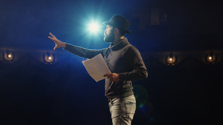 Mężczyzna w kapeluszu stoi na scenie w teatrze. Reflektory są zwrócone na niego. W ręce trzyma scenariusz i przemawia.