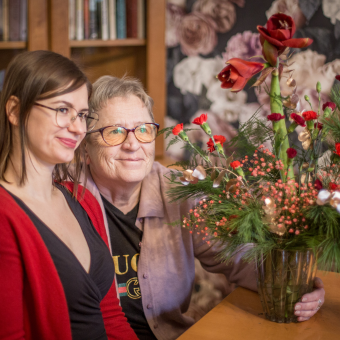 Seniorka i młoda kobieta wspólnie prezentują zrobiony przez nie stroik kwiatowy. 