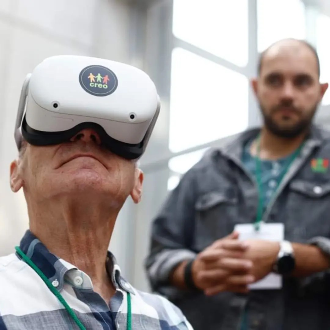 Senior zanurza się w wirtualnej rzeczywistości. Na głowie ma gogle VR. W tle jeden z trenerów CREO - Dawid.