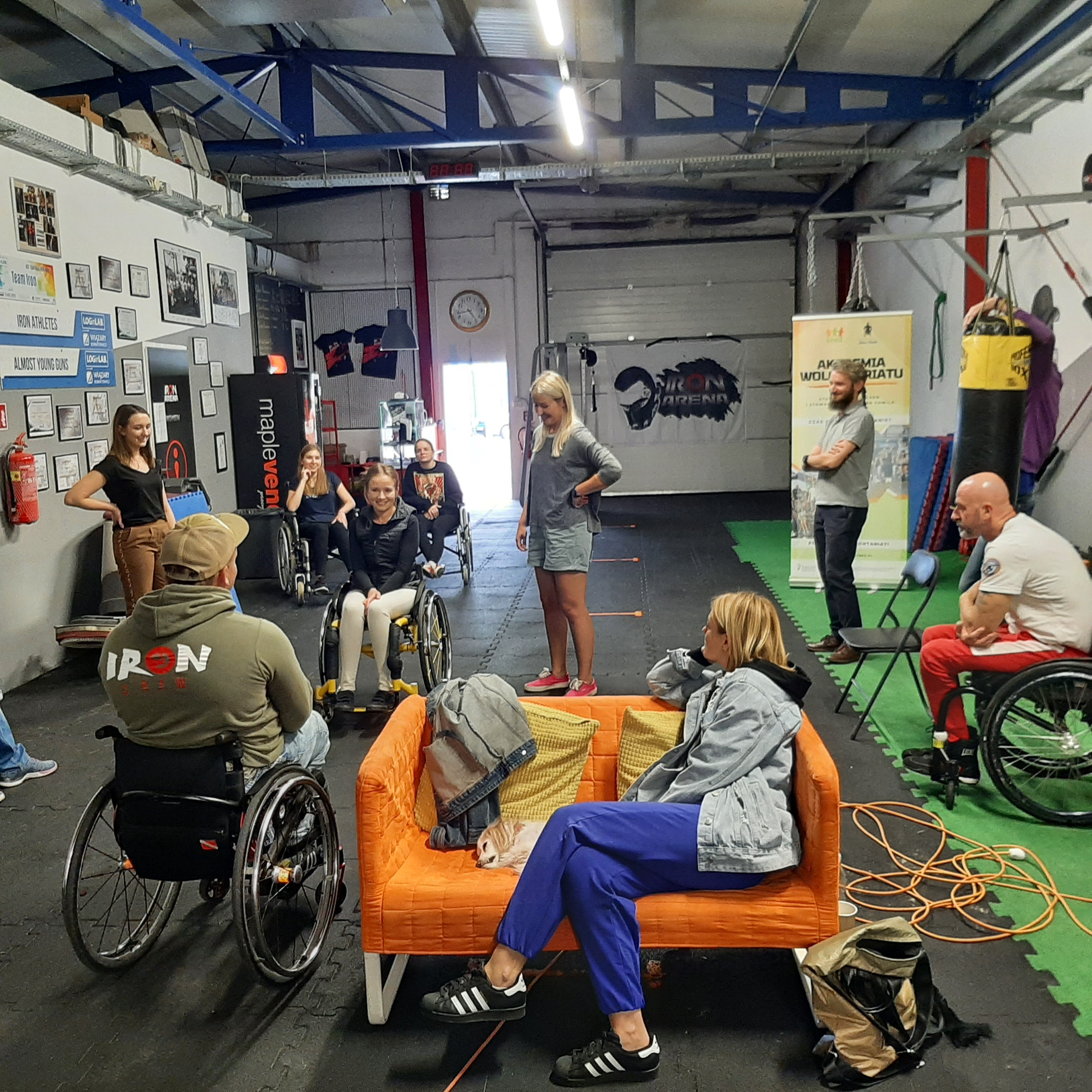 Grupa osób znajdująca się na sali crossfitowej. Część z nich porusza się na wózku. Wolontariusze trenują technikę jazdy wózkiem. Wszyscy są uśmiechnięci.