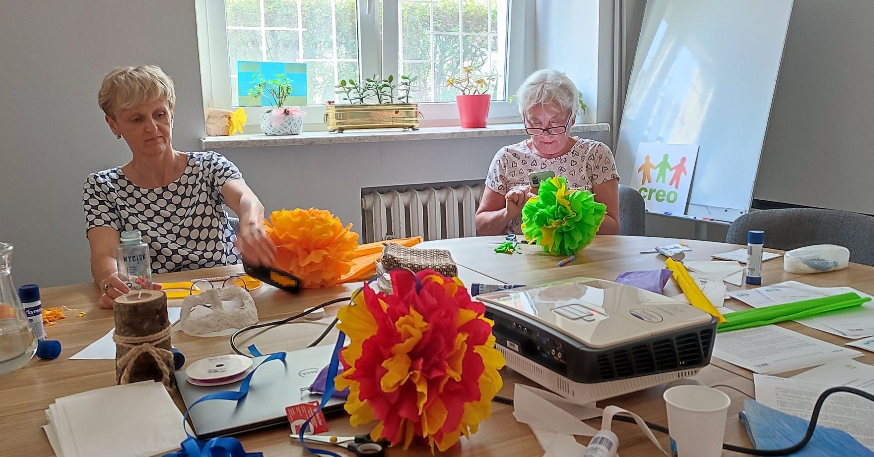 Dwie seniorki, siedzące przy stole. Robią kwiatki z bibuły. Na stole leżą różne artykuły papiernicze do tworzenia rękodzieła.